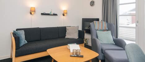 A modern living room with a sofa, armchair, and coffee table. There are wall-mounted lamps, a clock, and a television.