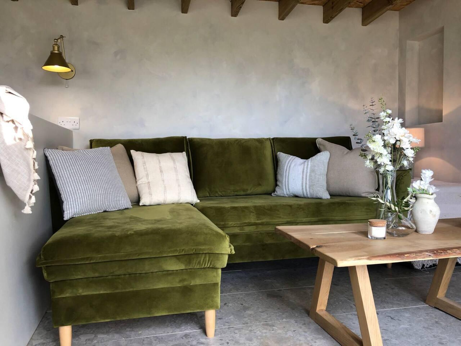 A green velvet sofa with multiple cushions, a wooden coffee table, and a vase with flowers.