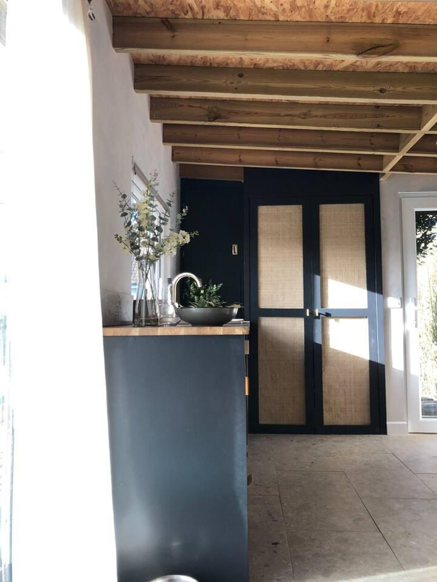 A modern kitchen with a dark blue door, a wooden ceiling, and a countertop with a vase of flowers.