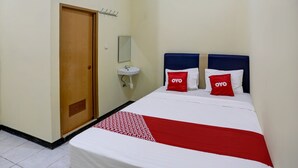 Habitación de hotel con puerta de madera, lavamanos blanco, espejo, cama con dos almohadas rojas y una colcha roja y blanca.
