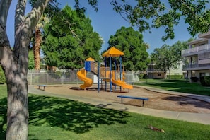 A playground with a yellow slide and a blue climbing structure, surrounded by trees and a fence. - Las Palmas Resort #2114 | All-Inclusive Amenities (Saint George)