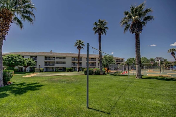 A well-maintained grassy area with a volleyball net, palm trees, and a building with multiple balconies in the background. - Las Palmas Resort #2114 | All-Inclusive Amenities (Saint George)