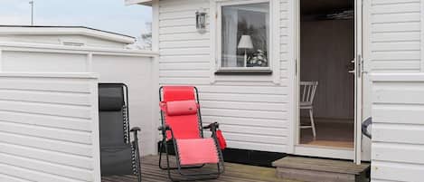 A small white mobile home with a covered deck, a red lounge chair, and a black chair.