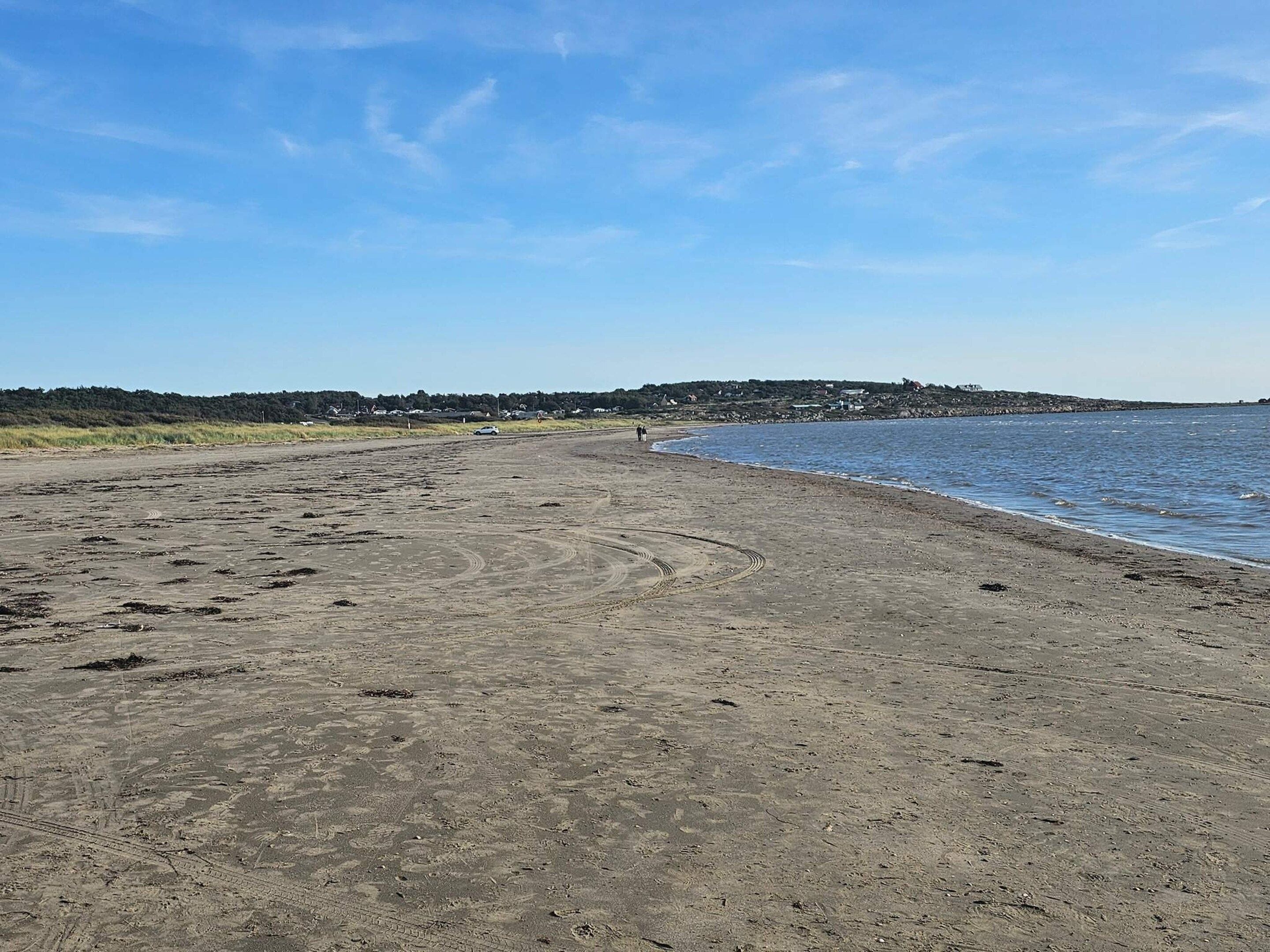 A sandy beach with footprints and tire tracks, a calm sea, and a distant shoreline with buildings.