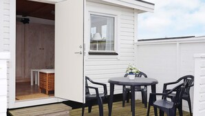 Een wit gebouw met een houten terras, een ronde tafel met stoelen en een raam met een witte gordijn.