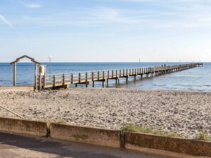 A beach with a wooden pier extending into the water, a lifeguard stand, and a concrete barrier separating the beach from the road. - 3 Person Holiday Home in Falkenberg (Falkenberg)