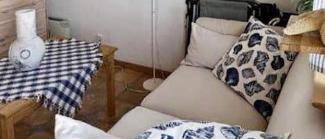 A room with a sofa, a small table with a checkered tablecloth, a fan, and a wall-mounted shelf with decorative items.