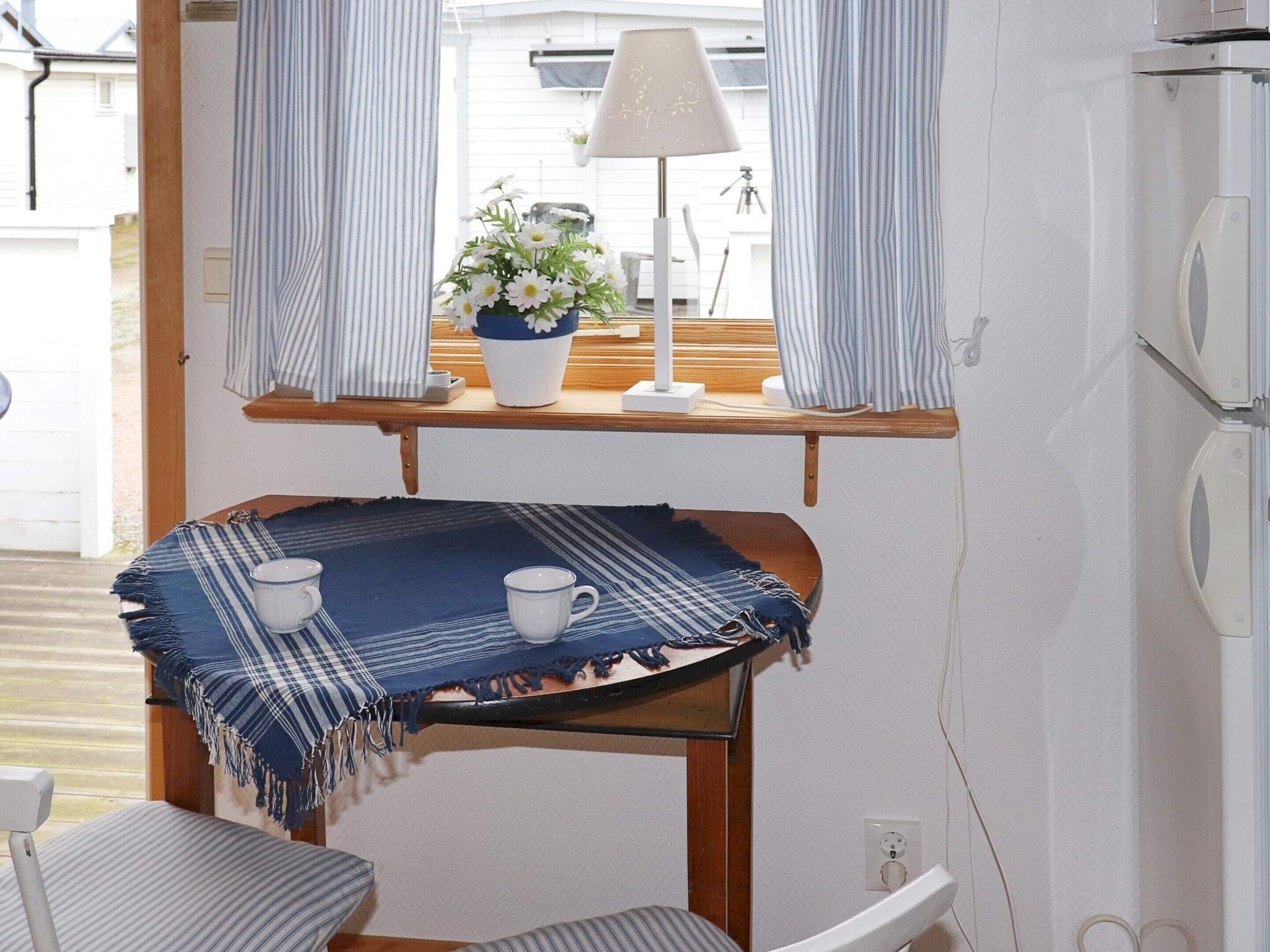 Una habitación pequeña con una mesa, dos sillas, una lámpara, una planta en maceta y un ventanal con cortinas.