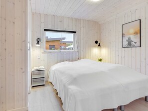 Un dormitorio con cama, ventana, mesita de noche, lámpara y un cuadro en la pared.