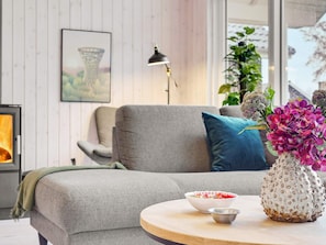A modern living room with a grey sofa, a round wooden table with bowls, a decorative vase with flowers, a lamp, and a fireplace. - 5 Star Holiday Home in Vaeggerlose (Vaeggerlose)