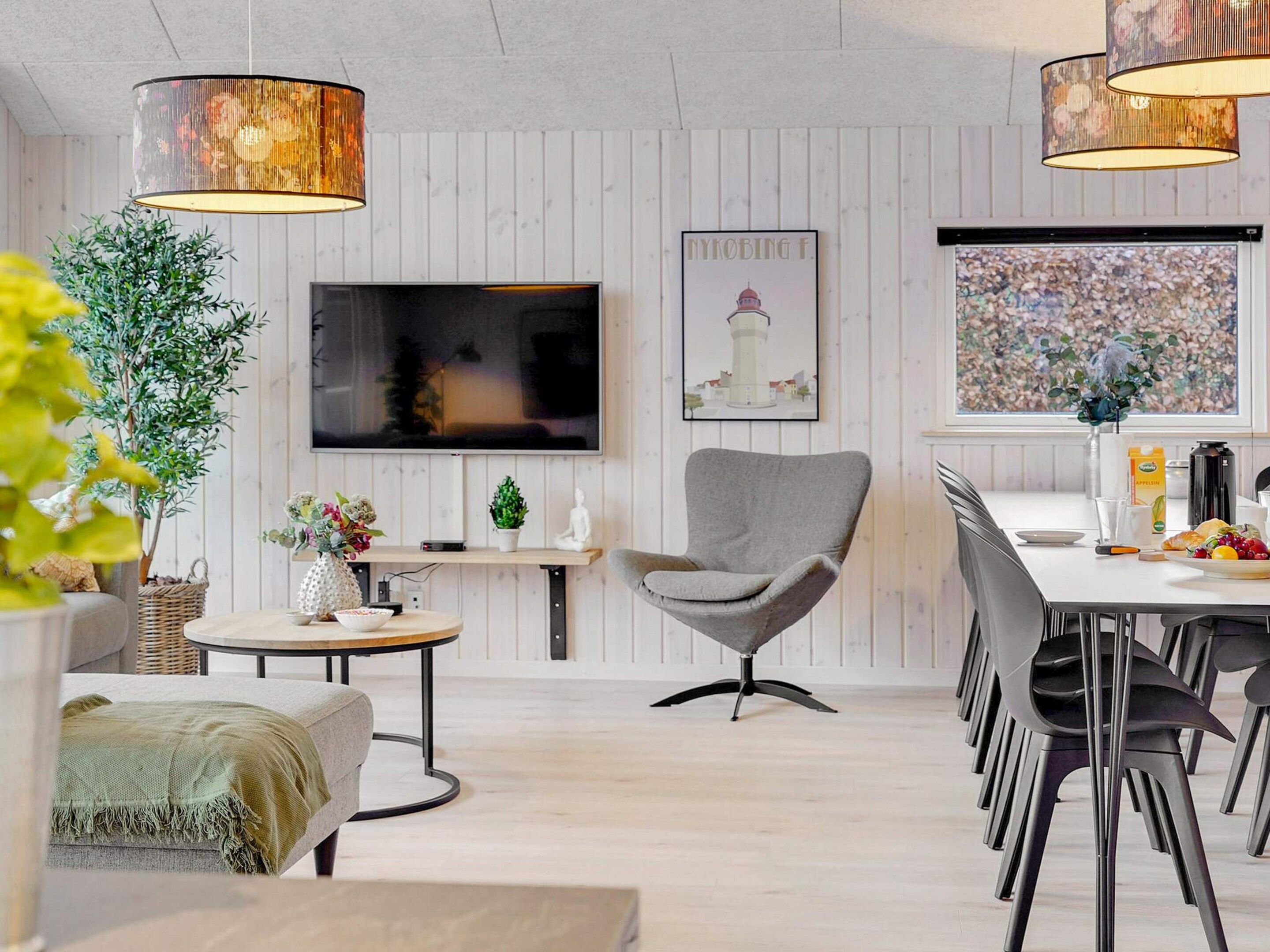 A modern living room with a flat-screen TV, a grey armchair, a dining area with a table and chairs, and a large window with a view.