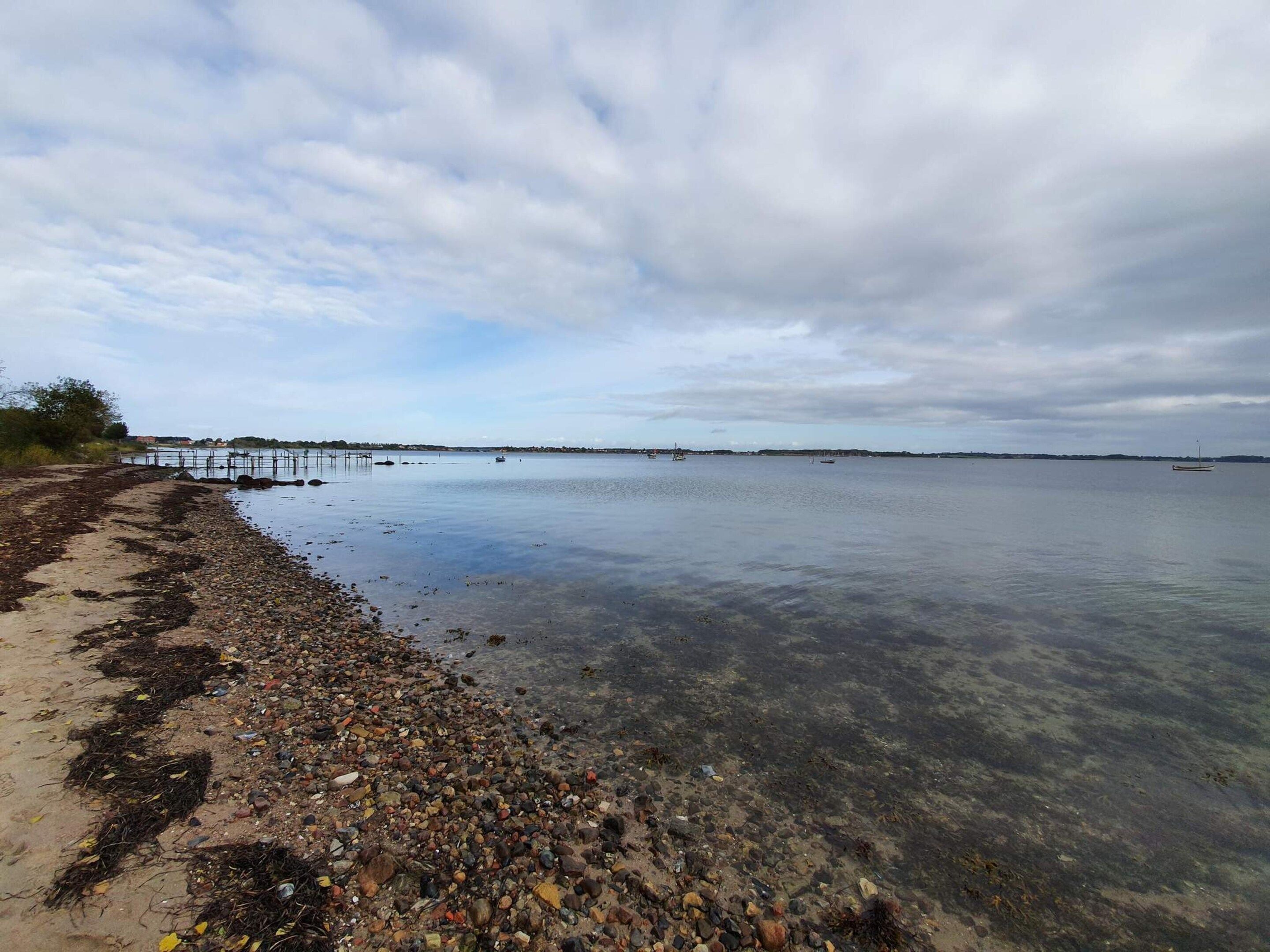 A beach with pebbles and seaweed, calm water, and a cloudy sky.