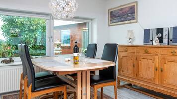 A dining area with a round table, chairs, a chandelier, a view of greenery outside, and a cabinet with decorative items.