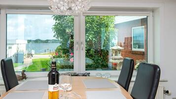 A dining table with a bottle of Black Tower wine, a glass, and placemats, overlooking a garden with a view of a lake and a brick building.