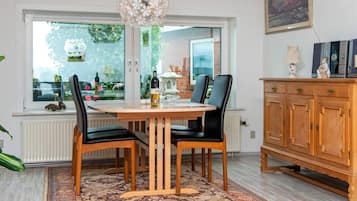 A dining area with a round table, two chairs, a patterned rug, a wooden cabinet, and a window with a view of greenery.