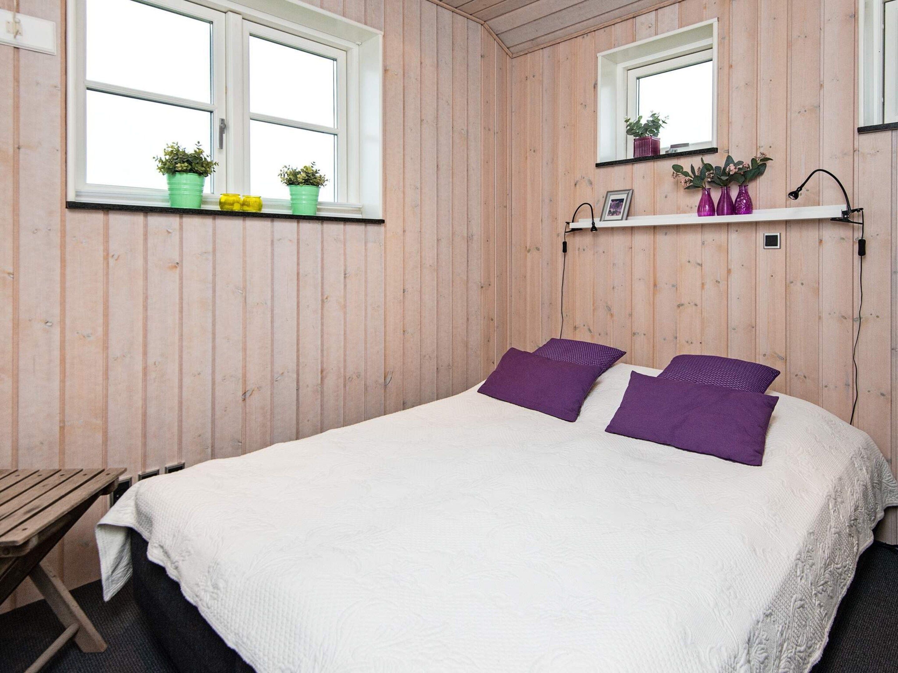 Een slaapkamer met een bed, twee ramen, een plank met planten en een bureau.