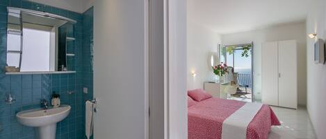 A modern hotel room with a bathroom featuring a sink and mirror, a bed with a red and white bedspread, and a balcony with a view.