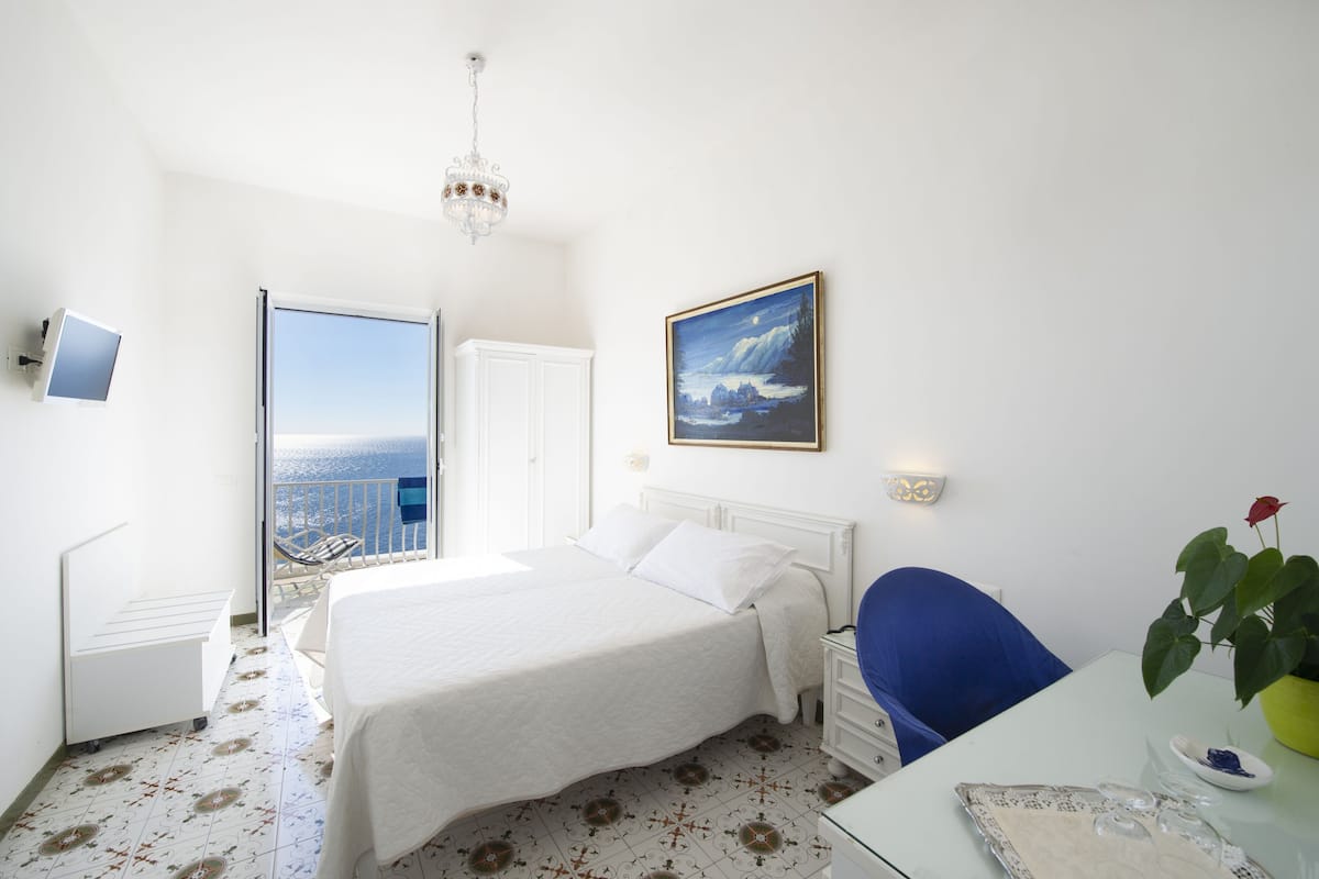 Ein Hotelzimmer mit Bett, Schreibtisch, Stuhl, einer Topfpflanze, einem Gemälde und Meerblick.