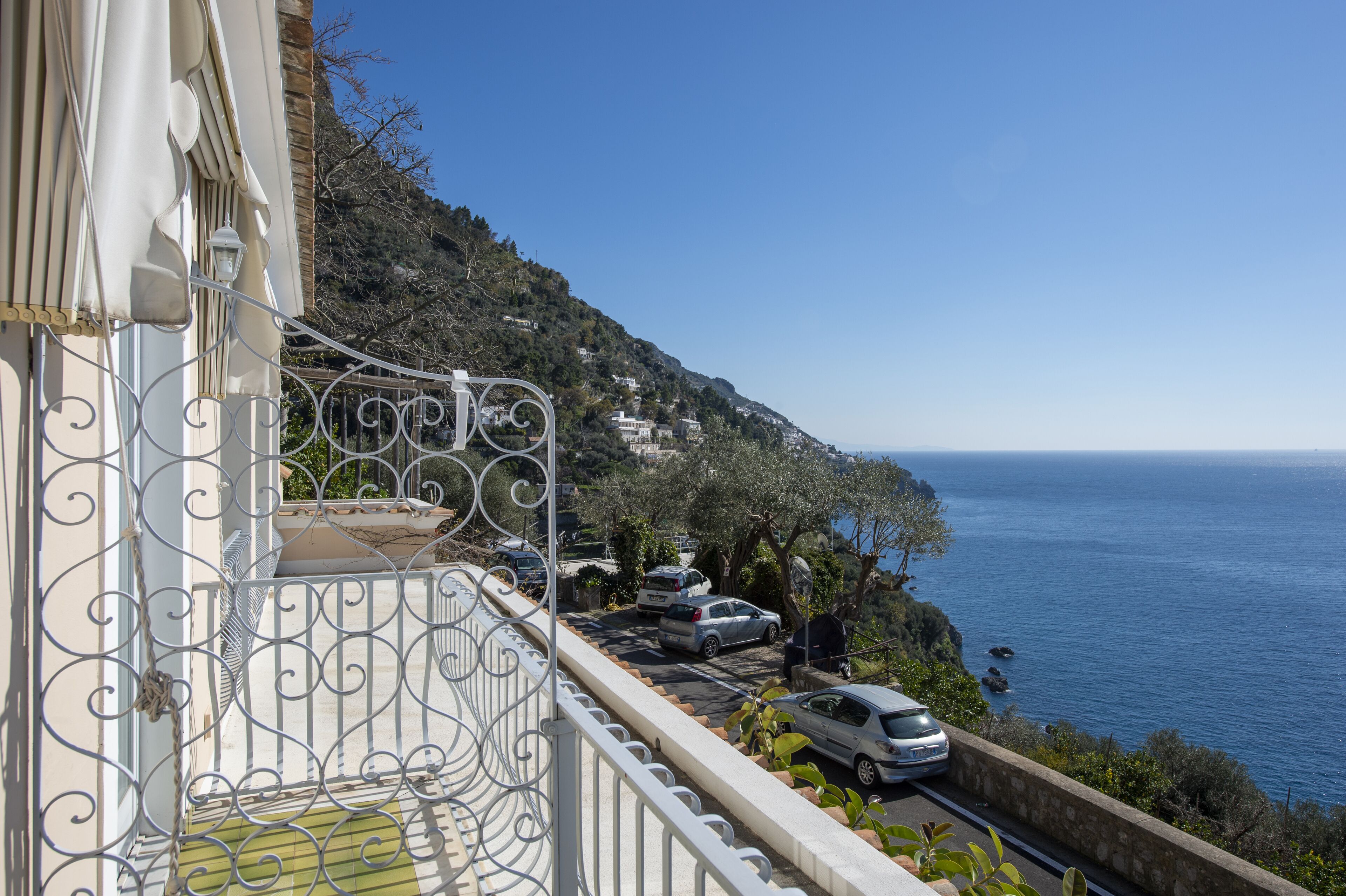 A balcony with a metal railing overlooking a coastal road with parked cars and a view of the sea.