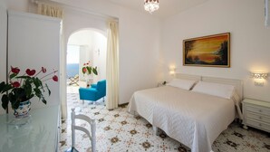 A bedroom with a bed, a nightstand, a painting, and a view of the sea through an open door. - Il Barilotto (Positano)