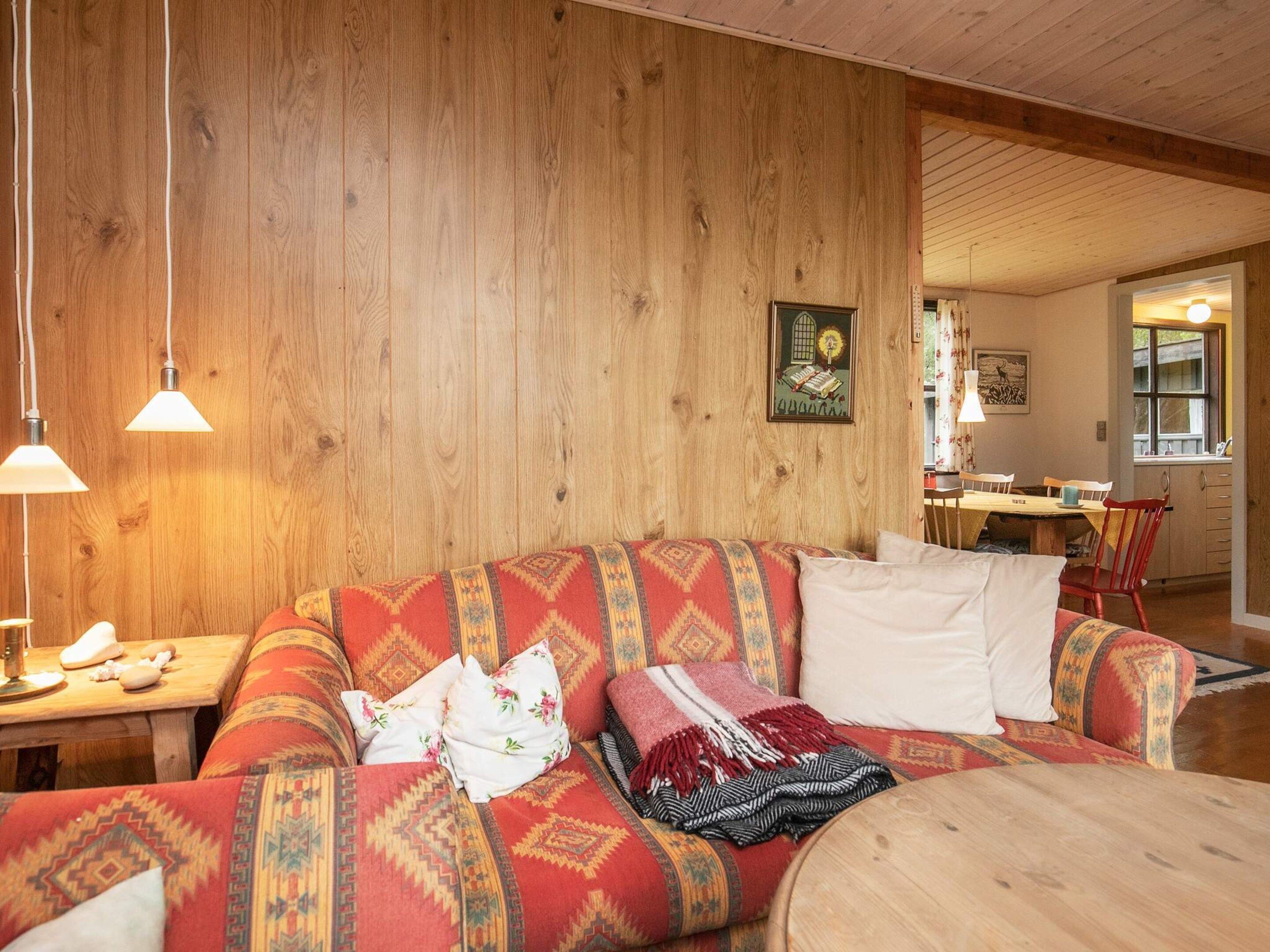 Una sala de estar acogedora con una pared de madera, un sofá con cojines estampados, una mesa de comedor con sillas y una cocina visible a través de una puerta abierta.