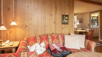 Una sala de estar acogedora con una pared de madera, un sofá con cojines estampados, una mesa de comedor con sillas y una cocina visible a través de una puerta abierta.
