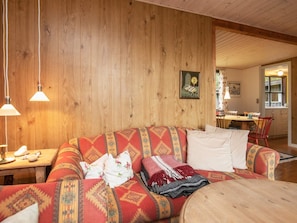 A cozy living room with a wooden wall, a sofa with patterned cushions, a dining table with chairs, and a kitchen area visible through an open door.