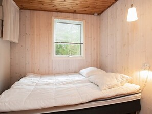 Un lit simple avec une literie blanche, dans une pièce aux murs en bois et dotée d’une fenêtre avec des stores.