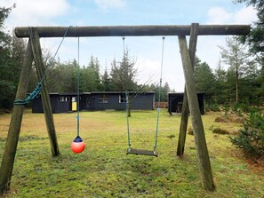 A wooden swing set with ropes and a red ball, set against a backdrop of a grassy yard and two small buildings.