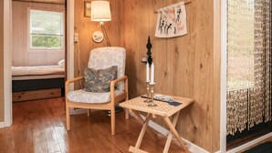 A cozy room with a wooden floor, a bed with white bedding, a patterned armchair with a cushion, a small table with a lamp, and a wall hanging.