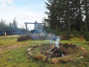 Een kampvuur omringd door rotsen en gras, met een bebost gebied en een gebouw op de achtergrond.