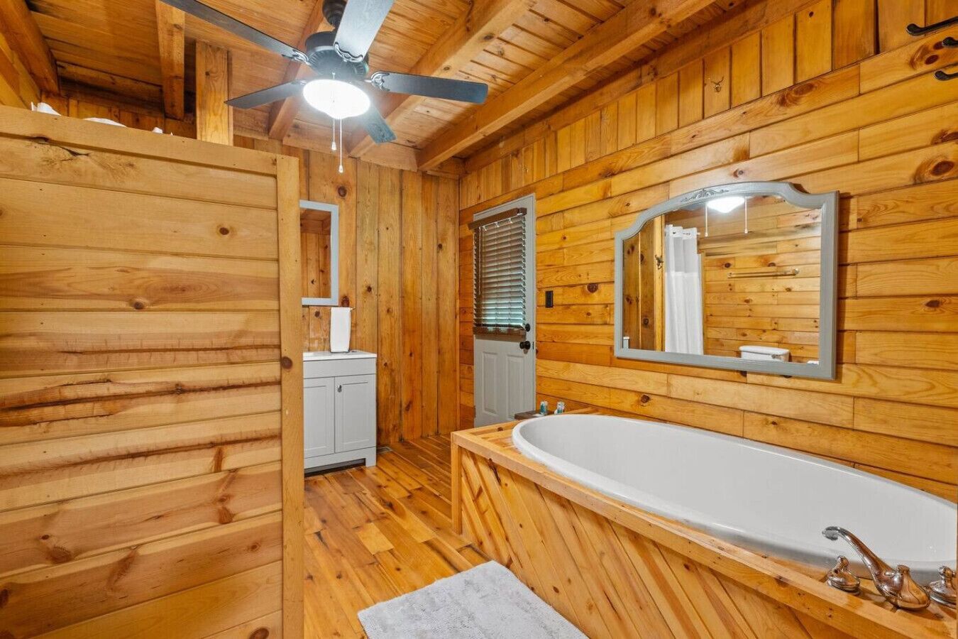 Een houten badkamertje in een chalet met een groot bad, een plafondventilator en een spiegel.
