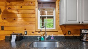 A kitchen with wooden walls, a granite countertop, a stainless steel sink, a toaster, a coffee maker, and a window with a view of greenery. - The Road`s End- Pet Friendly, Hot Tub, Firepit, Covered Deck (Pine Ridge)