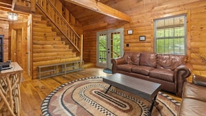 A log cabin interior with a staircase, leather sofa, and a patterned rug. - The Road`s End- Pet Friendly, Hot Tub, Firepit, Covered Deck (Pine Ridge)