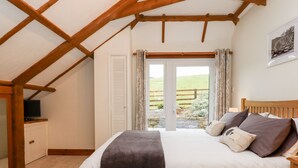 A bedroom with a sloped ceiling, a bed with pillows, a television on a cabinet, and a view of a green landscape through a window. - Little Orchard (Newton Abbot)