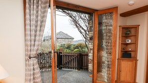 A room with a wooden floor, a sliding glass door leading to a garden, a small wooden cabinet, and a doormat. - Little Orchard (Newton Abbot)