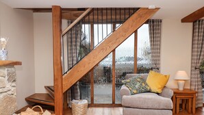 A cozy living room with a wooden staircase, a grey sofa with floral cushions, a wicker basket, and a small table with a lamp. - Little Orchard (Newton Abbot)