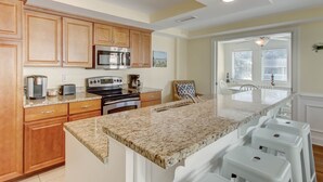 A kitchen with wooden cabinets, a stainless steel microwave, a gas stove, and a granite countertop. There are white bar stools and a view of the dining area through a glass door. - Rooftop Pool Access, Walk to Beach and Tybrisa (Tybee Island)