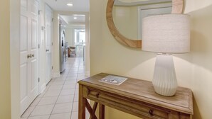 A hallway with a wooden console table, a white lamp, and a round mirror on the wall. - Rooftop Pool Access, Walk to Beach and Tybrisa (Tybee Island)