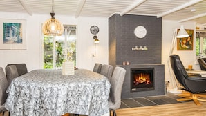 Ein modernes Wohnzimmer mit Kamin, einem runden Tisch mit Stühlen und einem großen Fenster mit Blick auf Grünflächen.