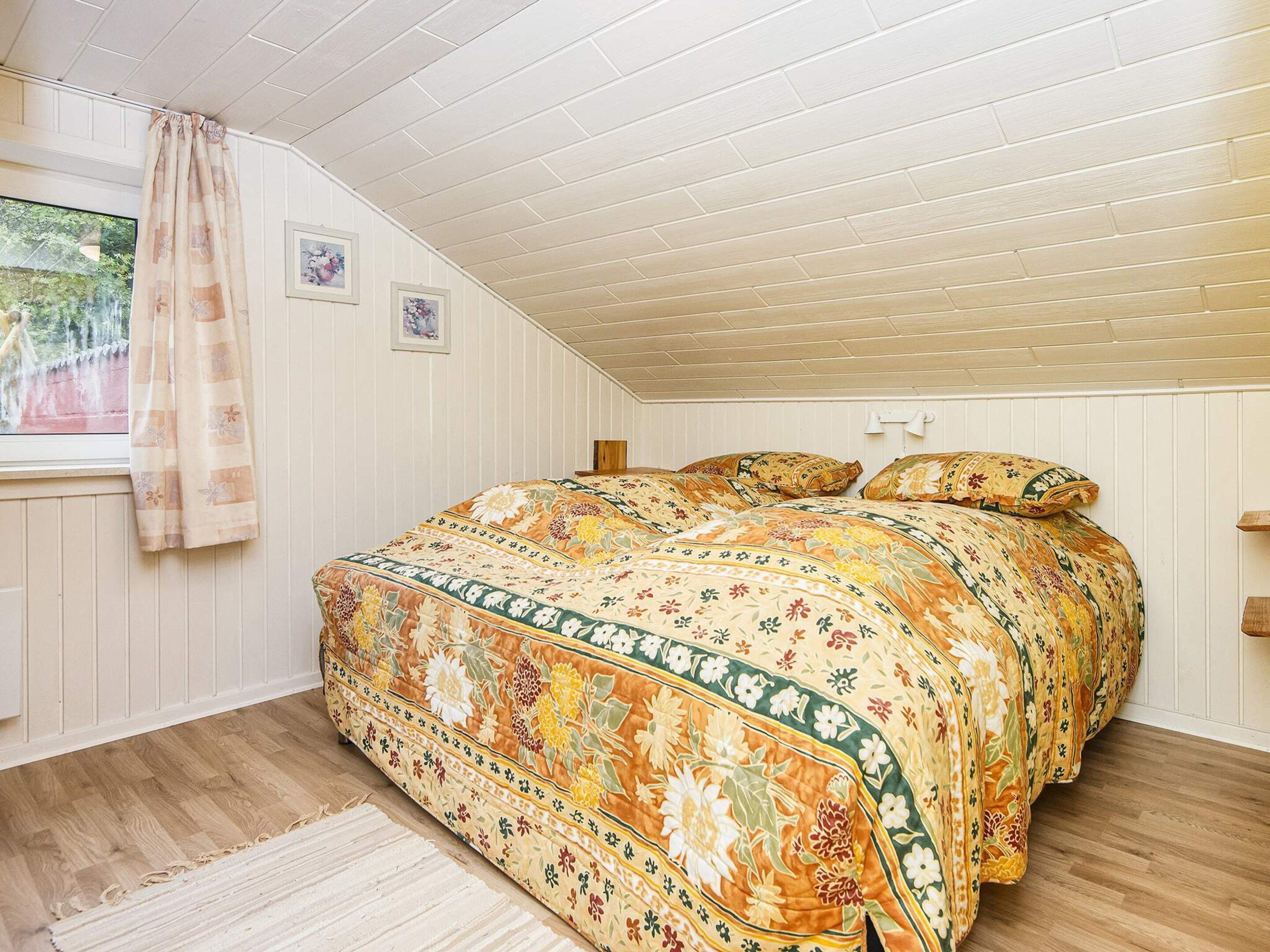 Un dormitorio con techo inclinado, suelo de madera, edredón estampado con motivos florales y una ventana con cortinas.