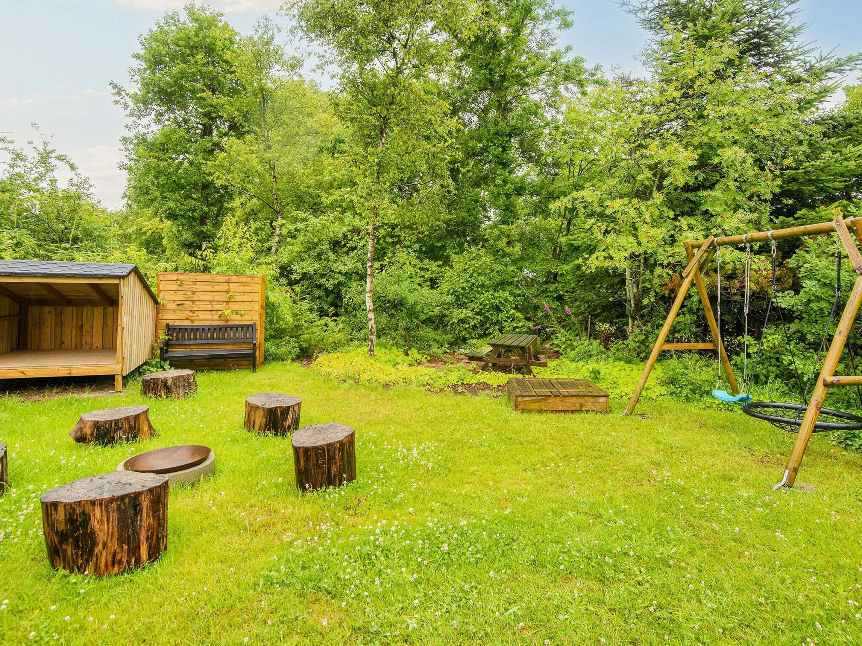 A wooden cabin with a bench and a swing set in a grassy area surrounded by trees.