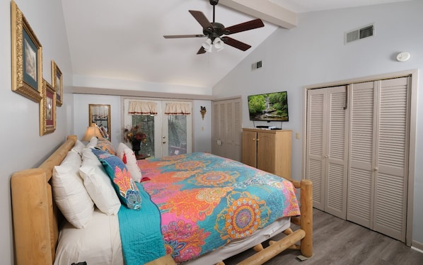 A bedroom with a bed, wooden headboard, ceiling fan, wardrobe, and a television on a cabinet.