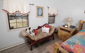 A room with a floral-patterned sofa, a wooden nightstand with a lamp, a bed with a colorful bedspread, and a window with curtains.