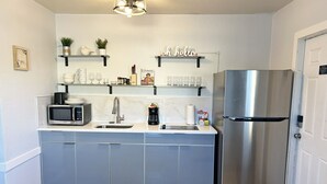 A compact kitchen with a stainless steel refrigerator, a microwave, and a sink. There are shelves above the counter holding various kitchen items and a 'oh hello' sign. - Private 1/1 Poolside Cottage! BBQ Grills, Near Beach & More! (Fort Myers)