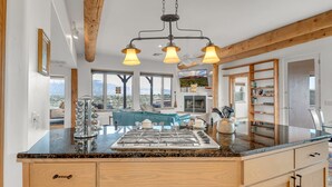 Una cocina con encimera de granito, armarios de madera y estufa integrada. Hay una zona de comedor con un sofá azul y un televisor sobre la chimenea.
