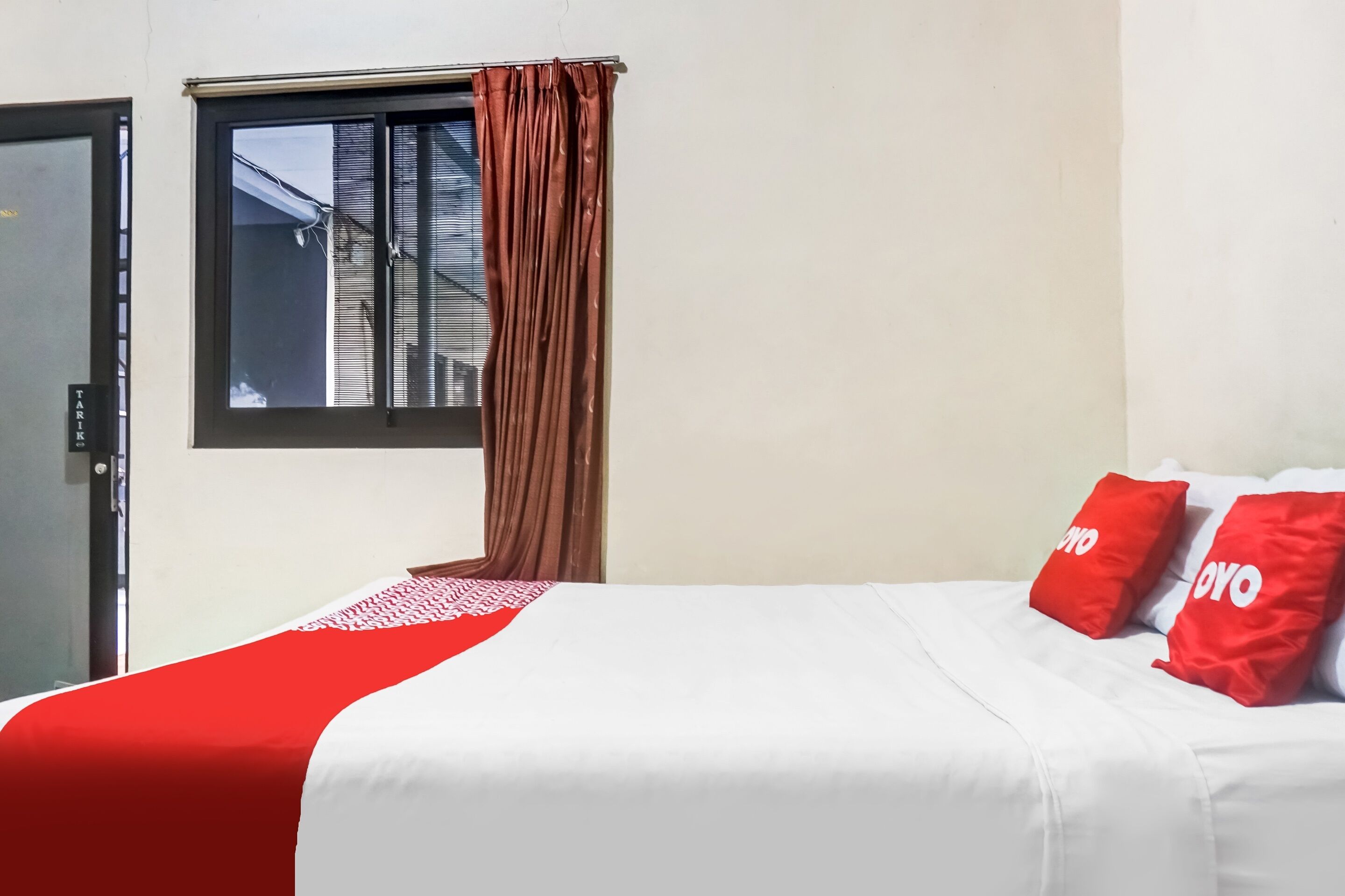 A hotel room with a bed, red and white pillows, a window with brown curtains, and a view of buildings outside.