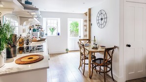 A bright kitchen with a dining area, wooden chairs, a clock, and a view of greenery outside. - Cozy Cotswold Dog Friendly Retreat  (Northleach)