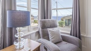 Ein Zimmer mit grauem Sessel, einem Holztisch und einer Lampe. Große Fenster mit Vorhängen bieten Blick auf das Meer und die Stadt.