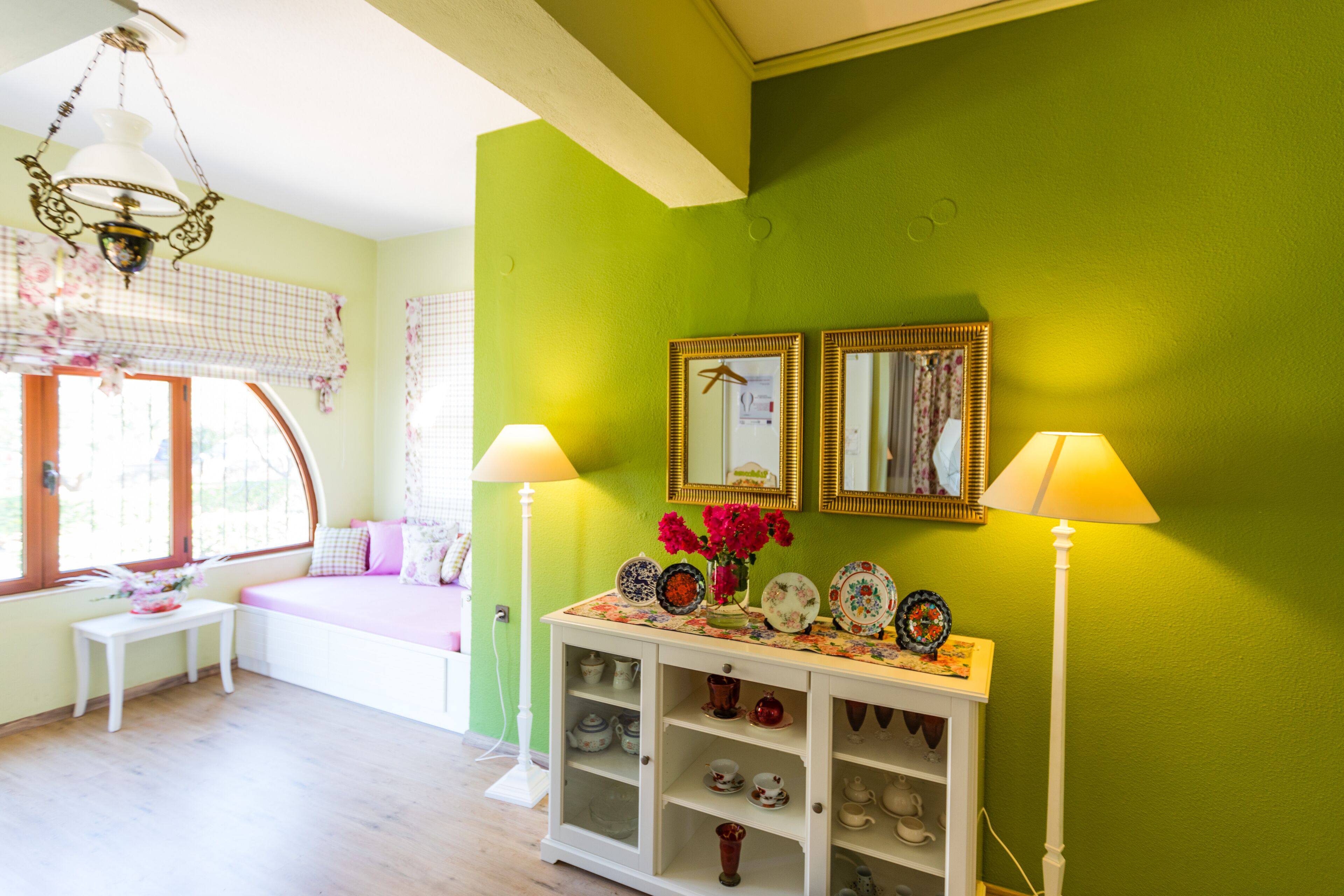 Ein Zimmer mit grüner Wand, einem weißen Beistelltisch mit dekorativen Tellern, einer Bank mit rosafarbenen Kissen und zwei Lampen.